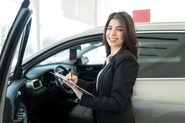 A woman next to a car holding a pen and notepad, symbolising the importance of timing when it comes to car insurance.